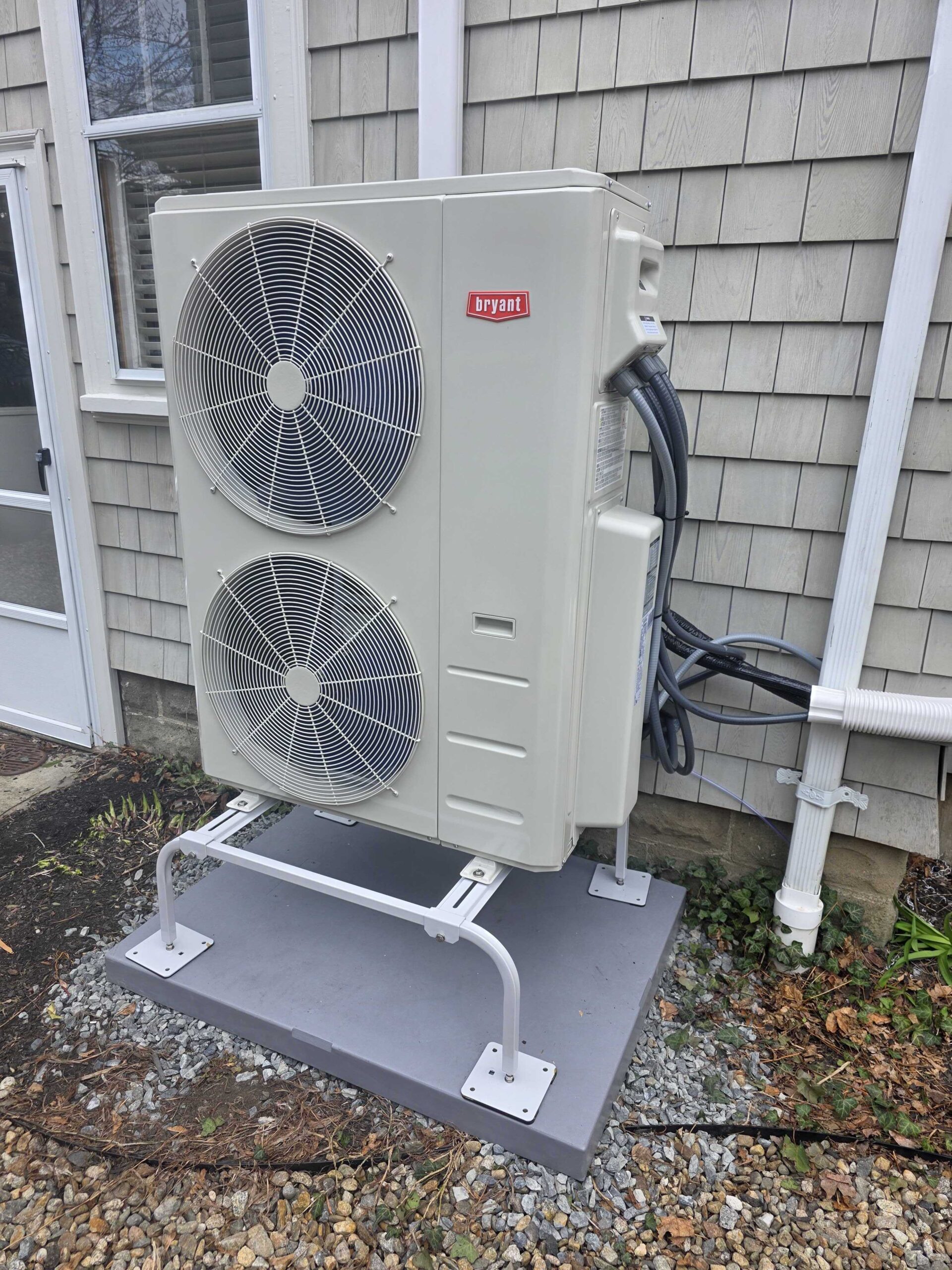 Outdoor Bryant heat pump unit mounted on a metal stand next to a house with gray shingle siding, connected by insulated pipes and cables.