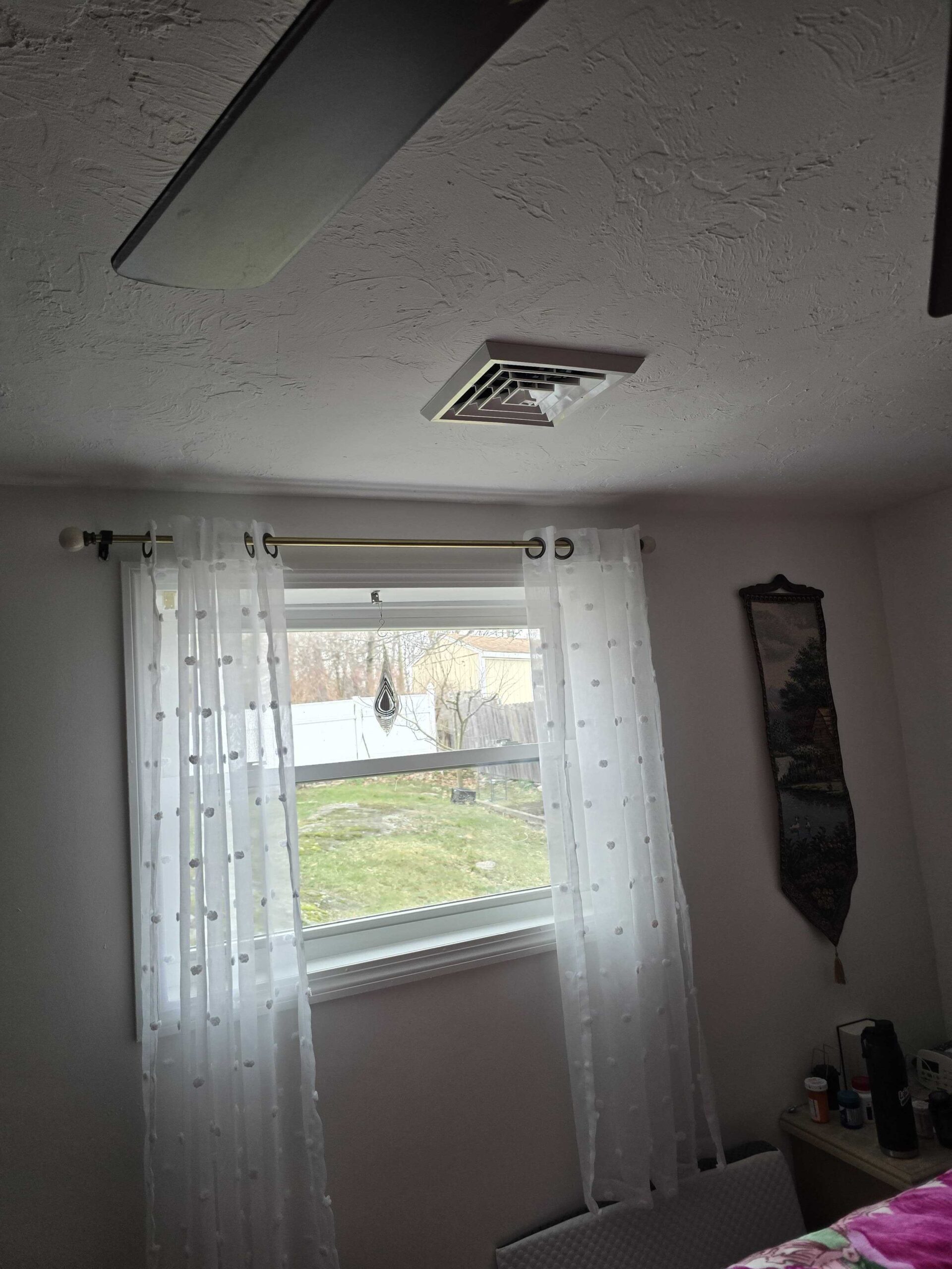 A window with sheer white curtains lets in natural light; a central AC supply vent and parts of a ceiling fan are visible above, with a yard scene outside.