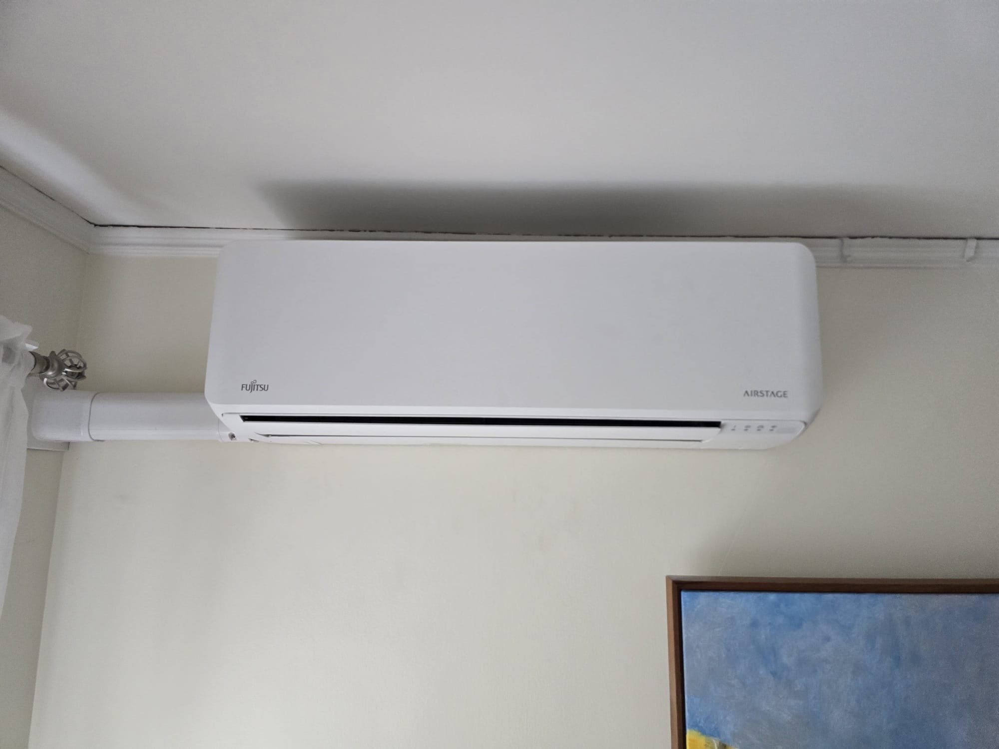 A white wall-mounted Fujitsu ductless minisplit unit installed near the ceiling, with part of a blue-framed painting visible in the bottom right corner.