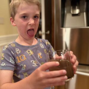 A child holds a glass of murky brown liquid, making a disgusted face and sticking out his tongue in a kitchen.