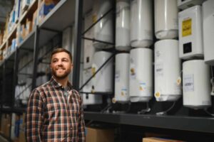A man in a plaid shirt stands in a warehouse aisle next to shelves filled with white water heaters.