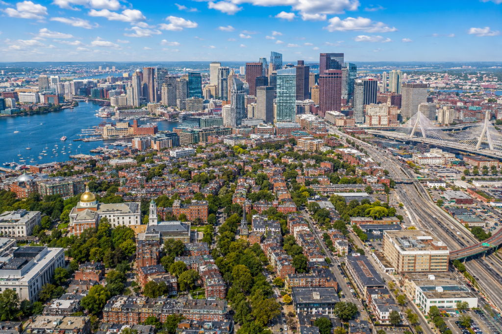 Aerial view of Boston, Massachusetts, featuring the downtown skyline, historic neighborhoods, Boston Harbor waterfront, major roadways, bridges, and urban green spaces under a bright blue sky.