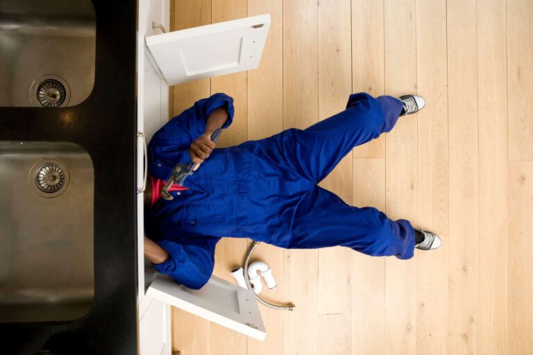 Person in a blue coverall lies on the floor under a kitchen sink, using a wrench to work on plumbing, with cabinet doors open and pipes nearby.