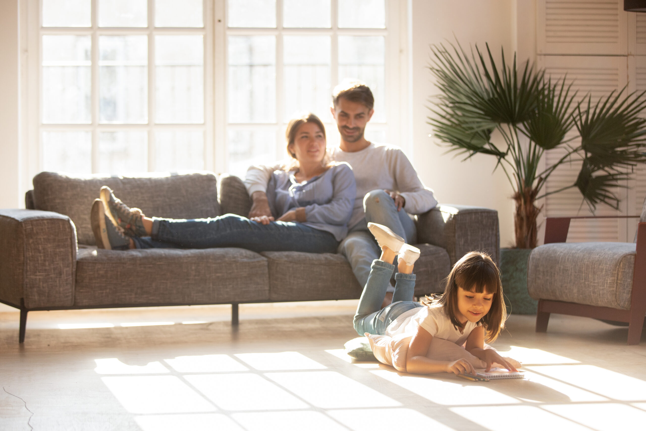 Happy parents relaxing on couch in comfort light living room while little kid child daughter playing on warm floor drawing with colored pencils