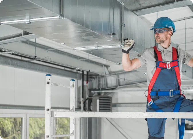 A construction worker in safety gear and a hard hat inspects or installs a metal ventilation duct in an industrial building.