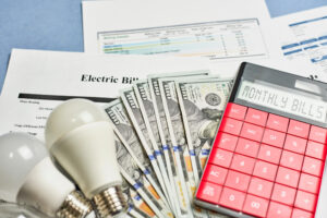 Image of electric bill displayed on a desk with a calculator, lightbulb, and a lot of cash representing increased home energy expenses in Massachusetts.