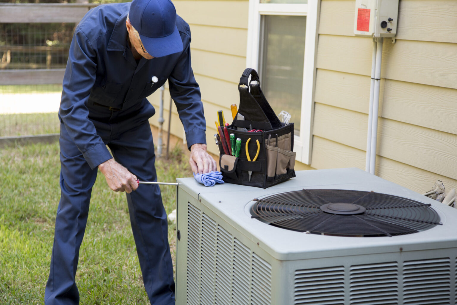 A technician in a blue uniform repairs an outdoor air conditioning unit using a screwdriver, with a tool bag placed on top of the unit.