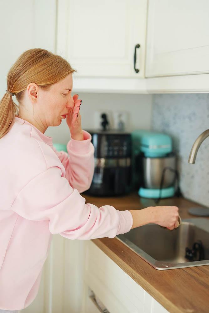 Woman reacting to unpleasant sewer smell while checking kitchen sink drain in home kitchen.